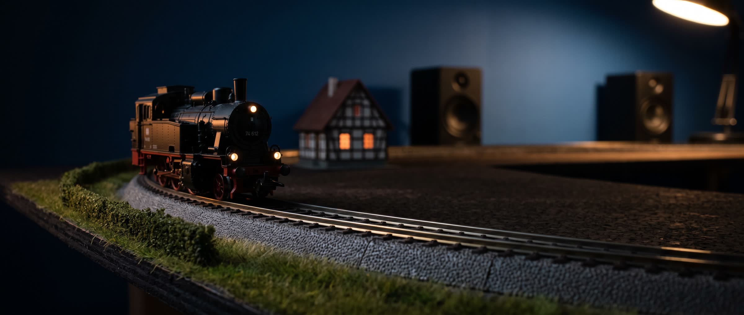 Eine Dampflok mit leuchtenden Lampen fährt am Abend an einem Fachwerkhaus mit erleuchteten Fenstern vorbei, dahinter zwei Lautsprecher